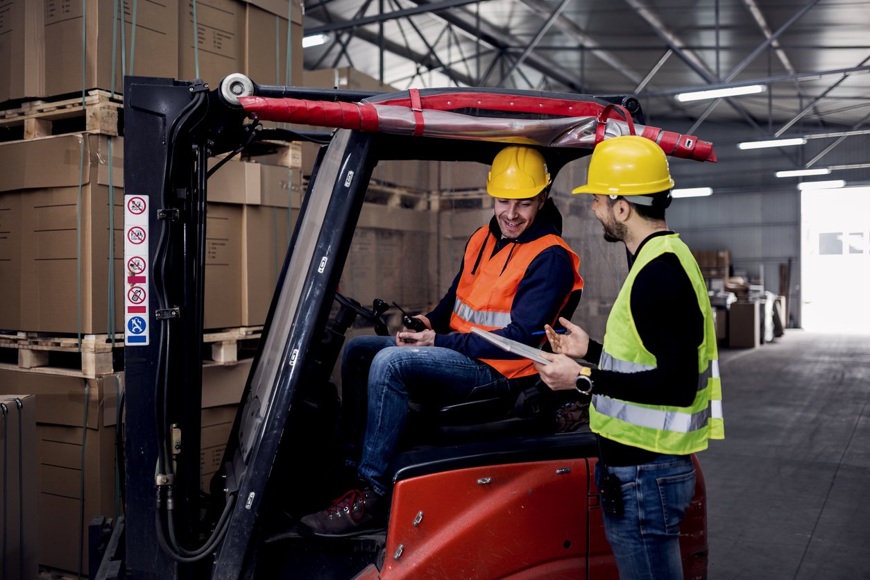 Twee mannen in een magazijn waarvan een op een kleine heftruck en de andere met een lijst. Op de achtergrond veel dozen op stapels met pallets.