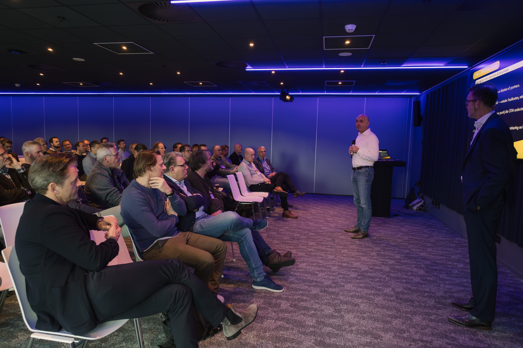 Bijeenkomst Open Trip Model met een grote groep mensen in het publiek en twee sprekers voorraan